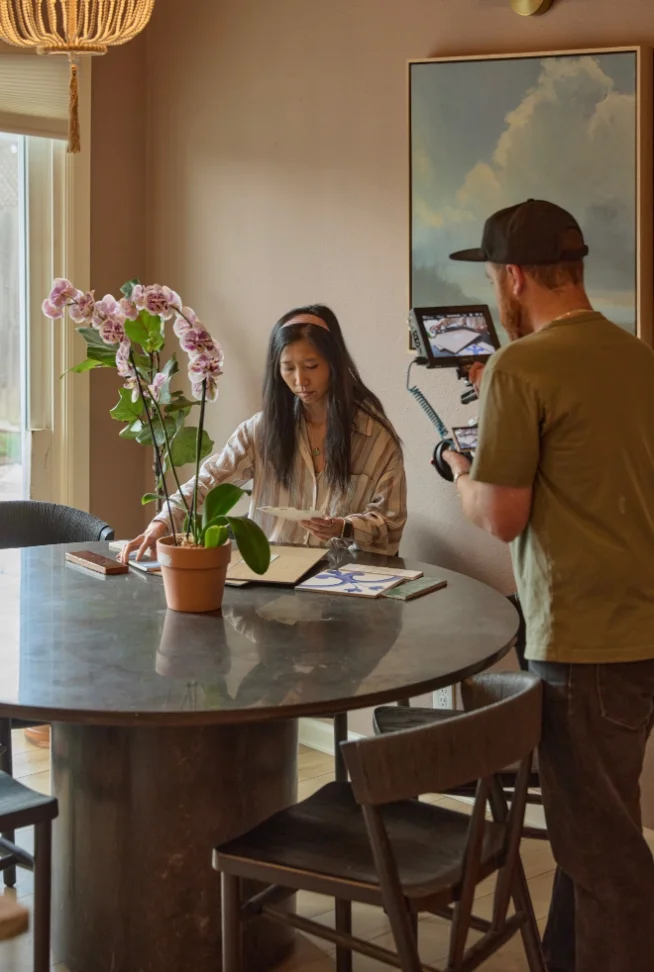 Florence reviewing plans and material samples at a round stone table with an orchid