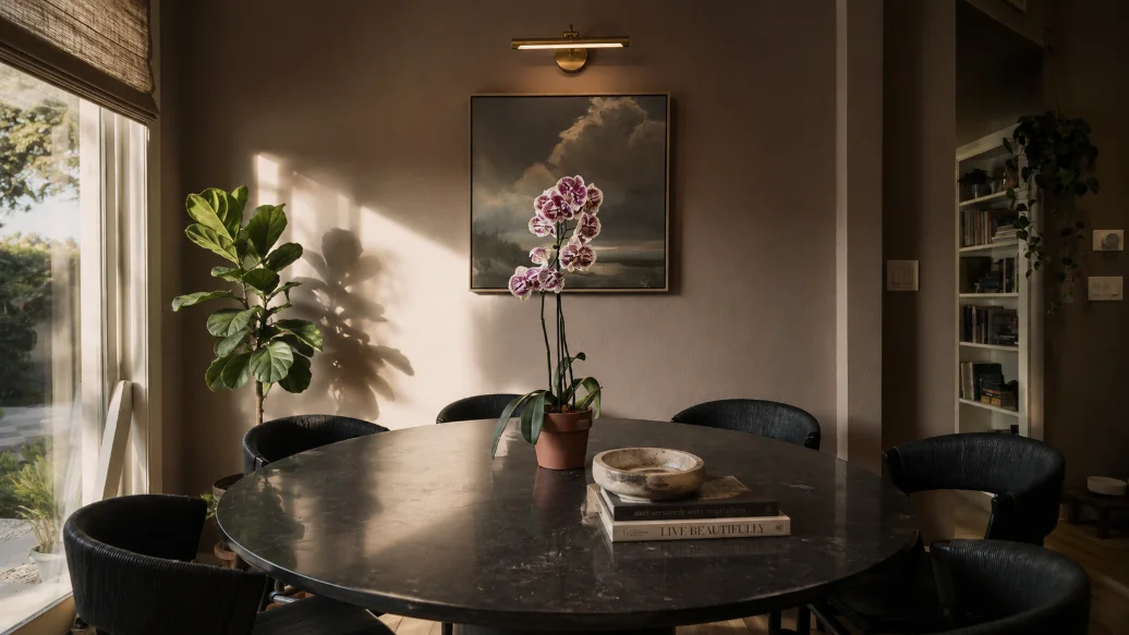 Dining room with a round black stone table, woven dark chairs and an orchid, lit by low evening sun
