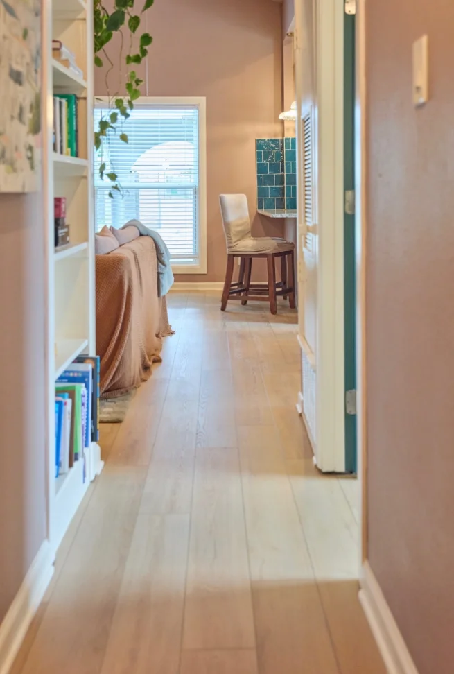Sunlit hallway with white oak floors, blush walls, a white bookcase and a teal-tiled nook